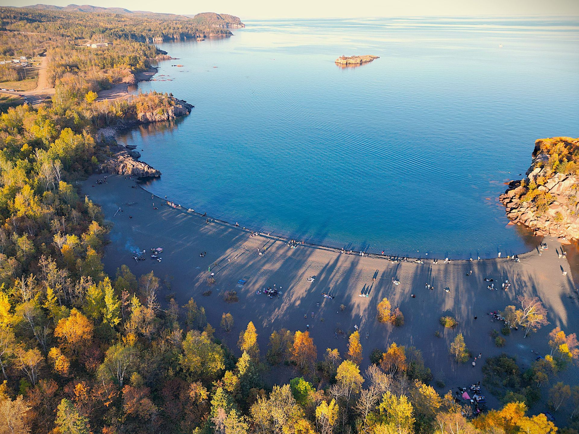 Aerial view of a bay with a dark sandy beach surrounded by trees in autumn colors, water, and a small island.