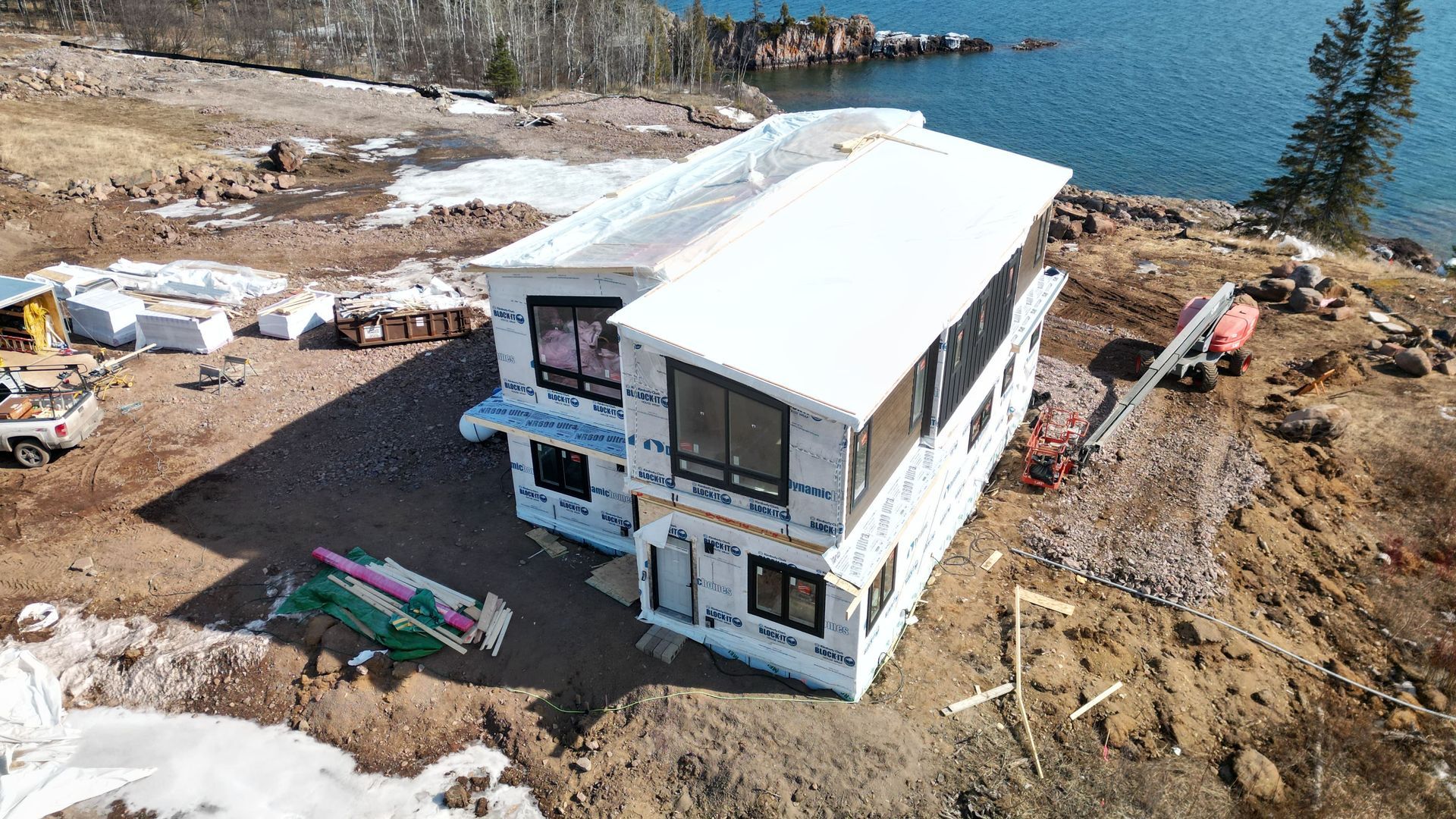 Two-story house under construction on a rocky shoreline with a lake in the background.