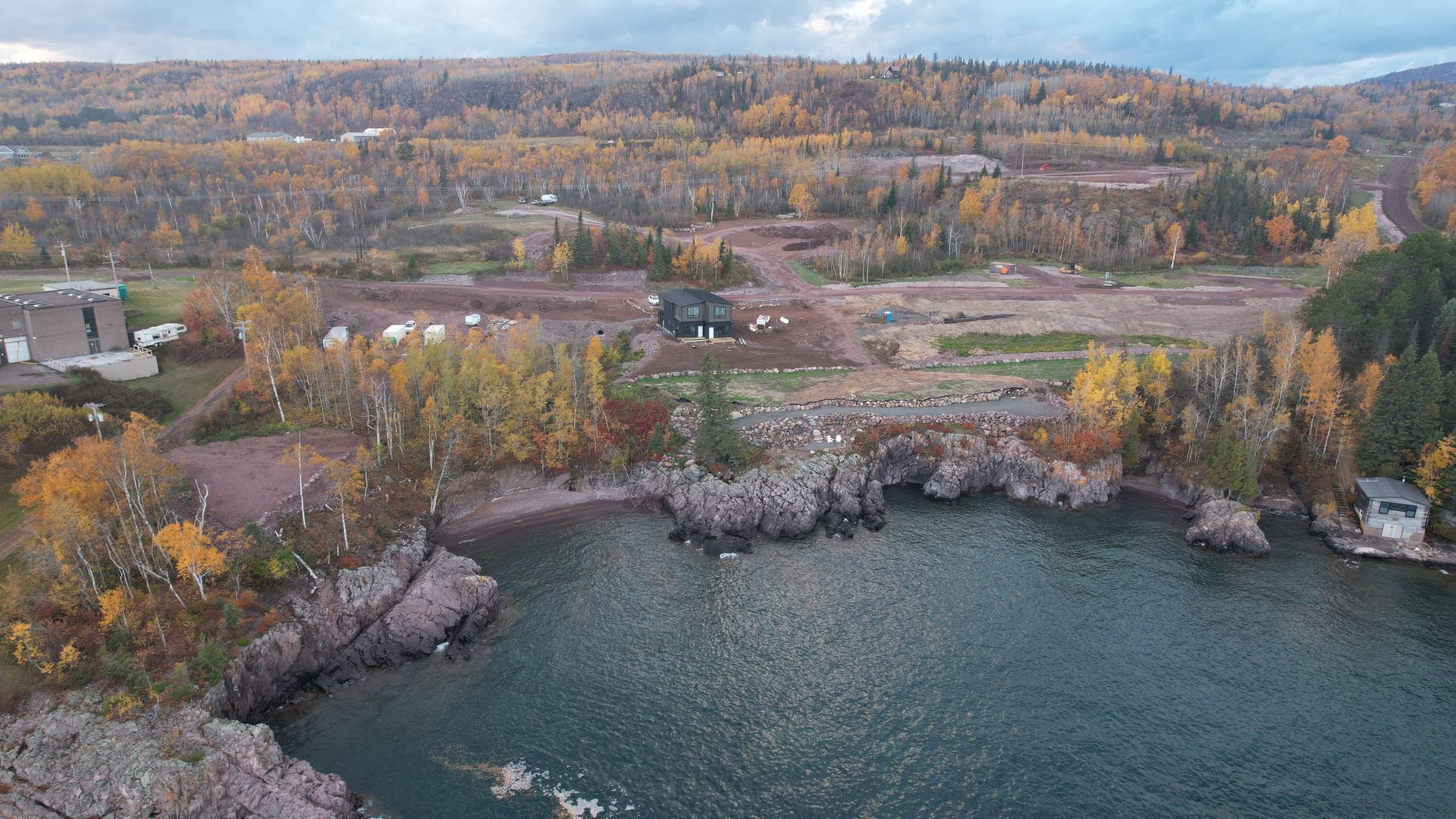 Coastal view of trees with fall foliage overlooking a lake and rocky shoreline; a house is on the land.