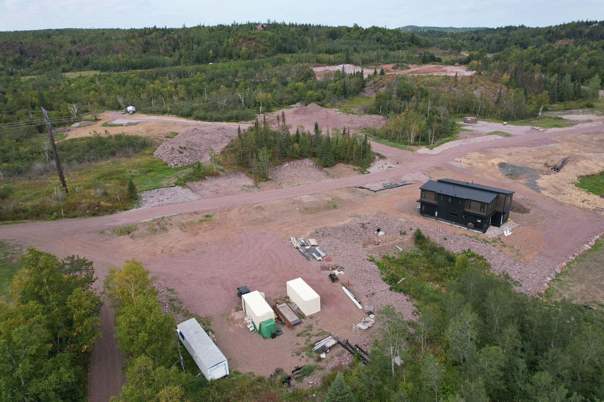 Aerial view: a construction site with a dark building, machinery, and gravel piles amidst green trees.