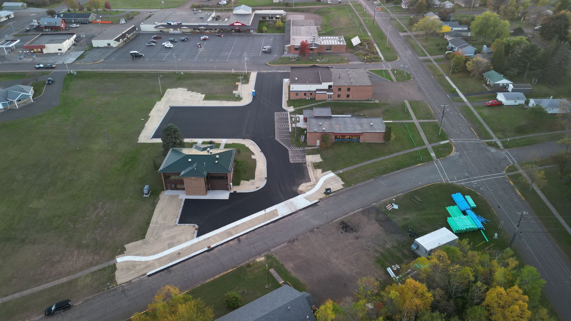 Aerial view of a town with buildings, roads, and a large parking lot. Green grass surrounds.