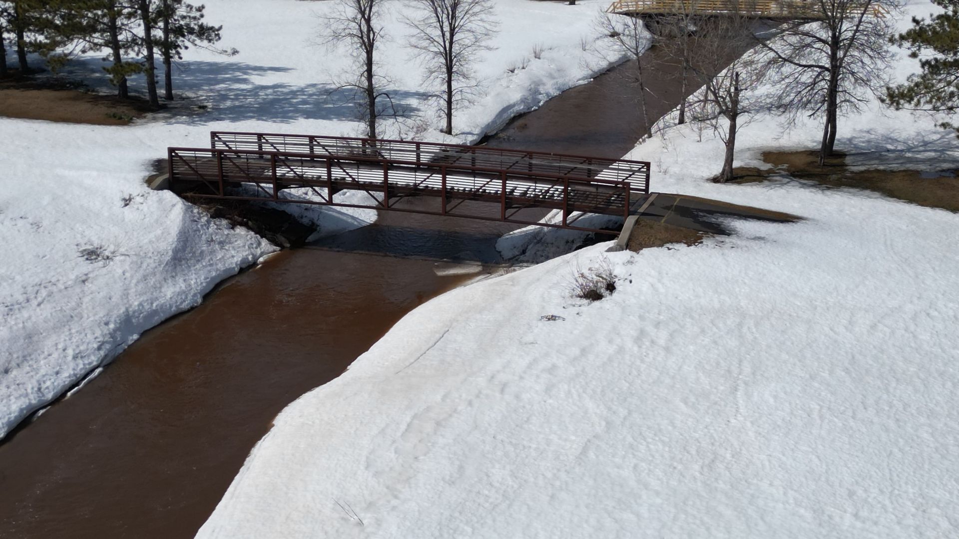 A wooden bridge over a brown stream in a snowy landscape. Trees line the banks.