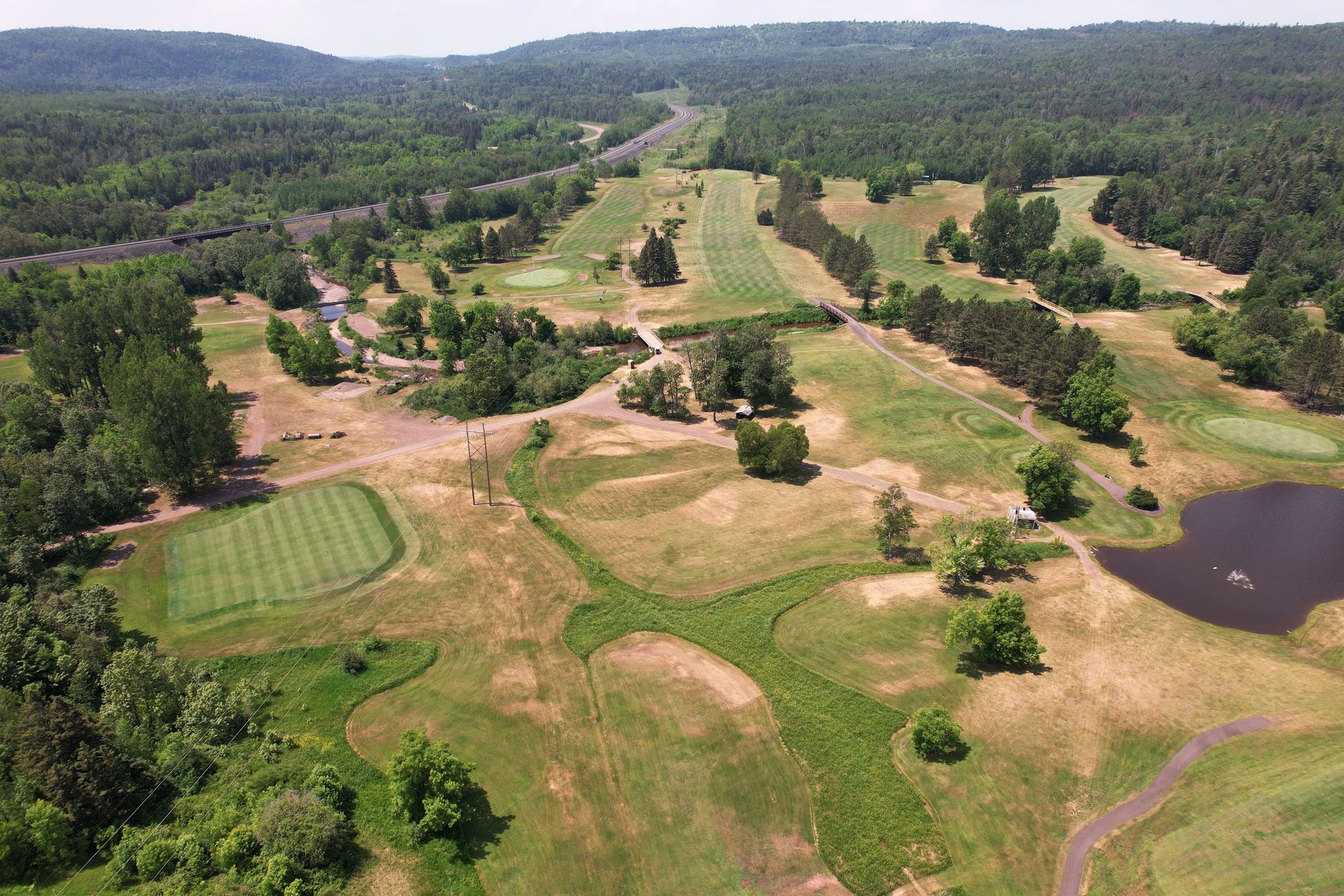 Aerial view of a golf course with green fairways, rough, trees, and a small pond. A train track is visible in the background.
