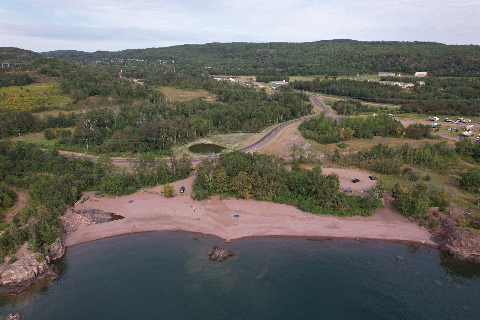 Aerial view of a sandy beach, surrounded by trees and a lake, with a road and buildings in the background.