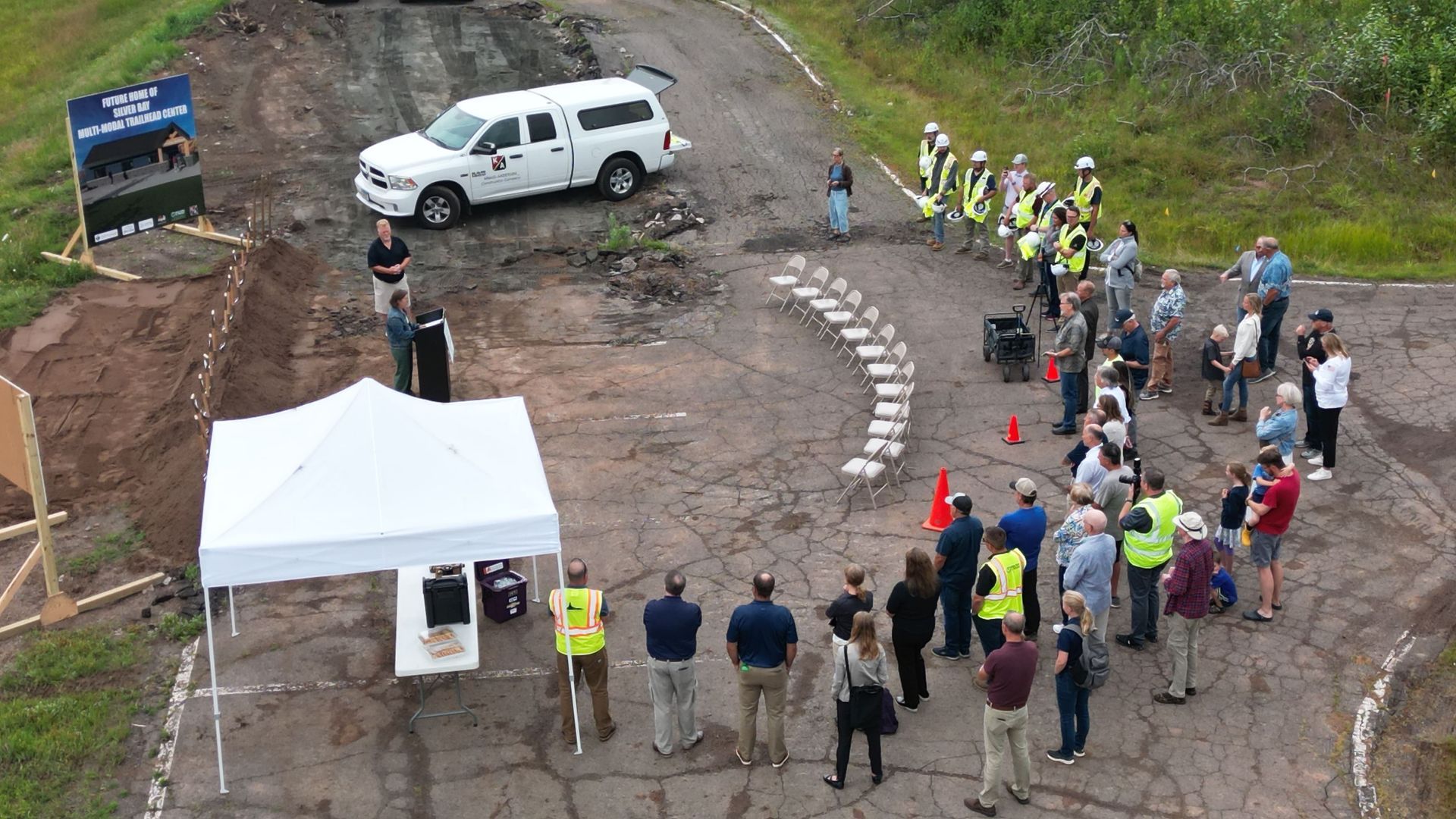 Groundbreaking ceremony: people gathered, white tent, truck, road work site.