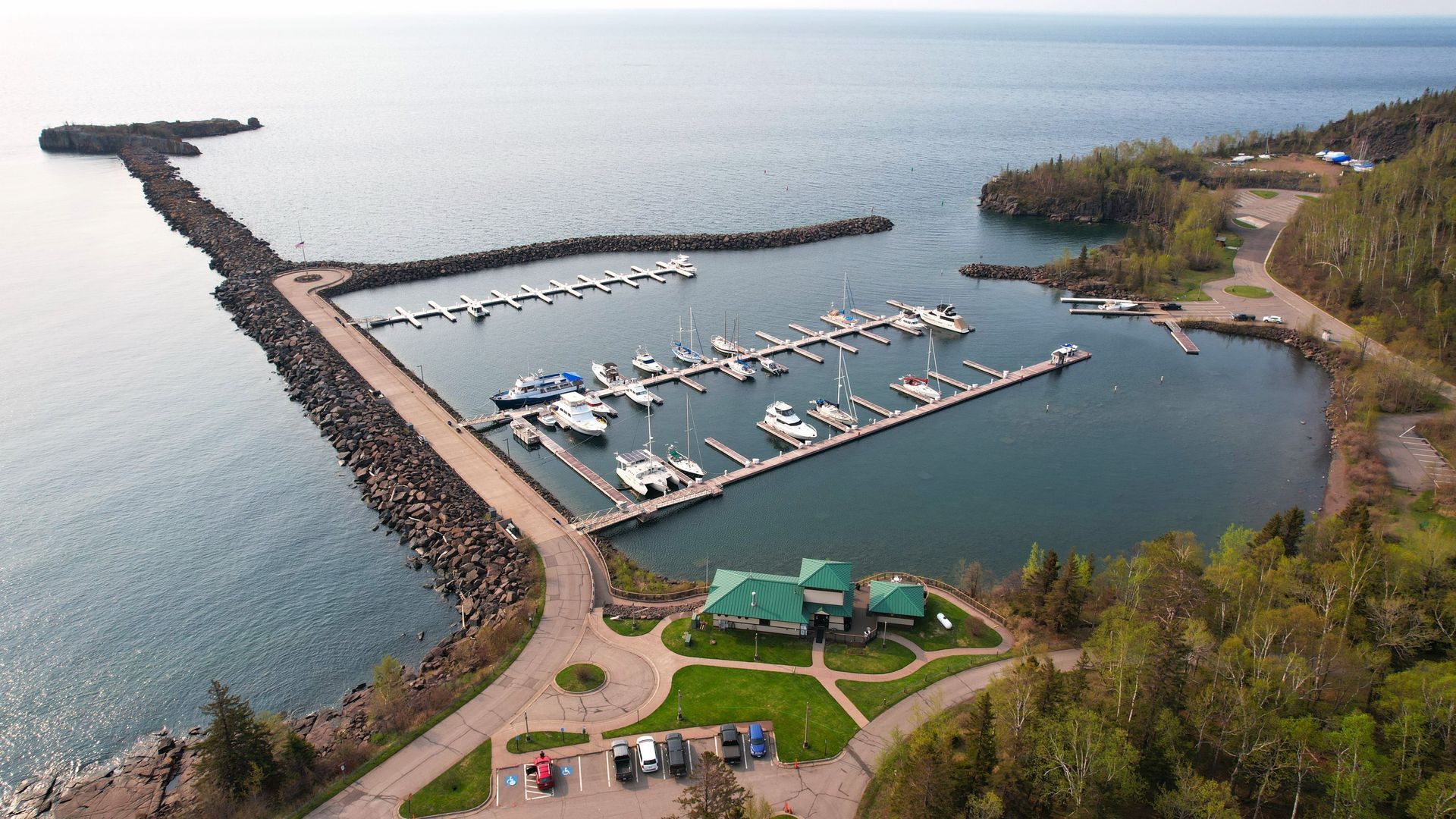 Marina with docked boats, surrounded by rocky breakwater, trees, and a building, on a lake.