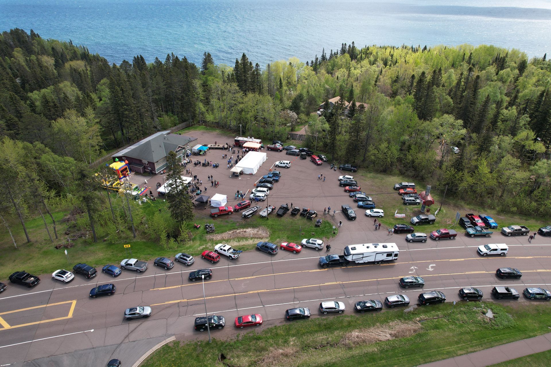 Aerial view of a car gathering near a building and forest, overlooking a lake. Numerous vehicles and people are present.