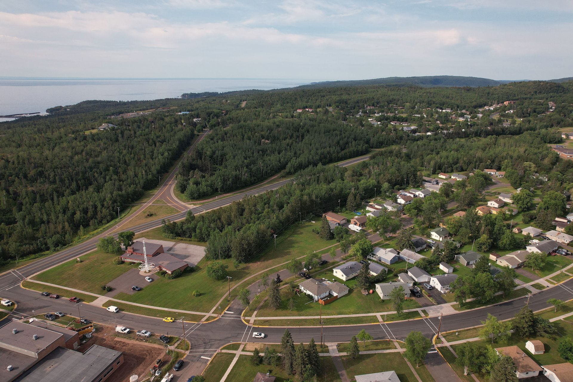 Aerial view of a town with houses, roads, and a forest next to a body of water under a blue sky.