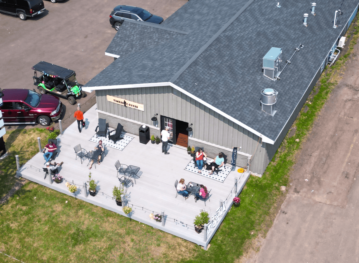 Aerial view of a gray building with a deck. People sit at tables. Cars and golf cart are parked nearby.