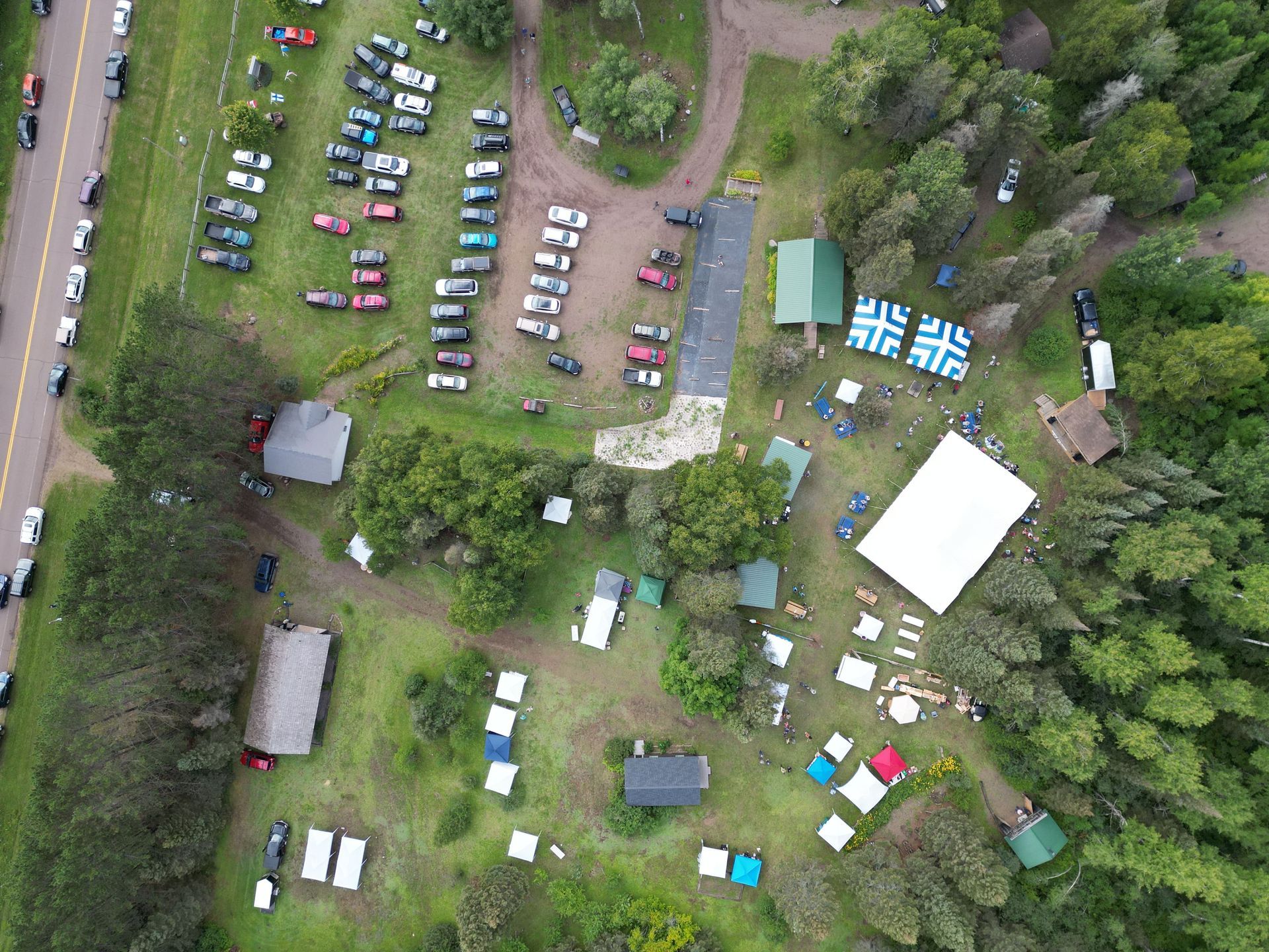 Aerial view of an outdoor event. Cars parked in a field, buildings, and tents among trees.