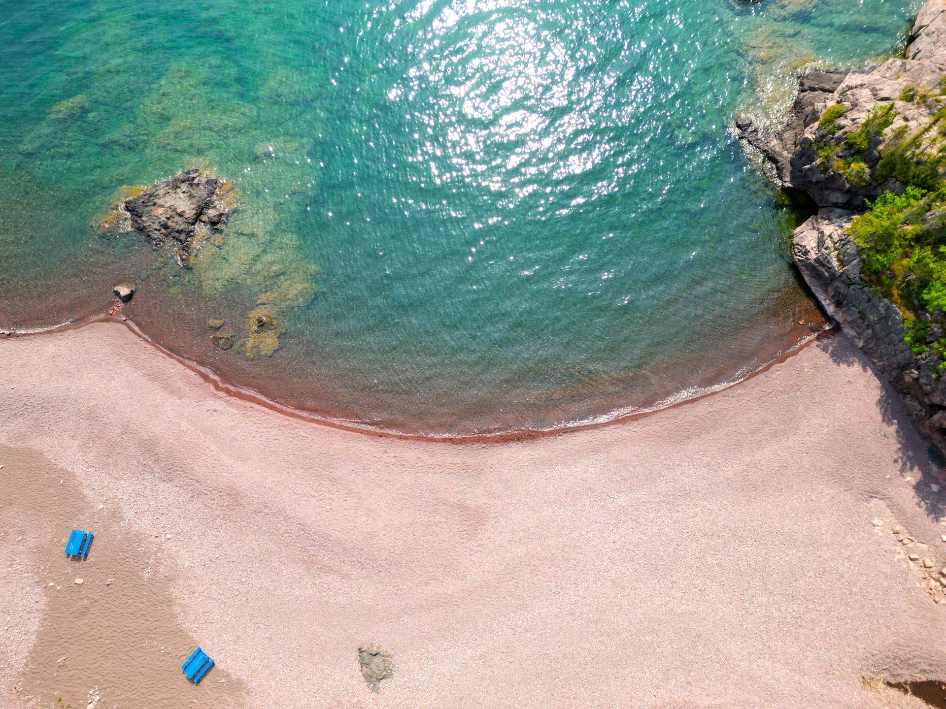 Aerial view of a pink pebble beach, turquoise water, and rocky cliffs.