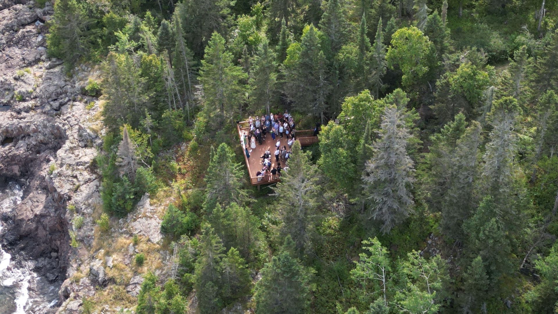 Wooden platform with people surrounded by trees, overlooking rocky coastline.