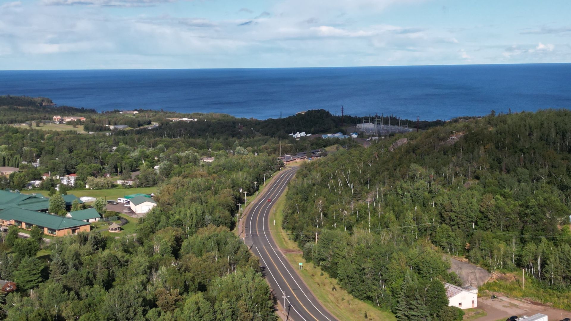 Curving road through green forest towards a blue lake under a cloudy sky.