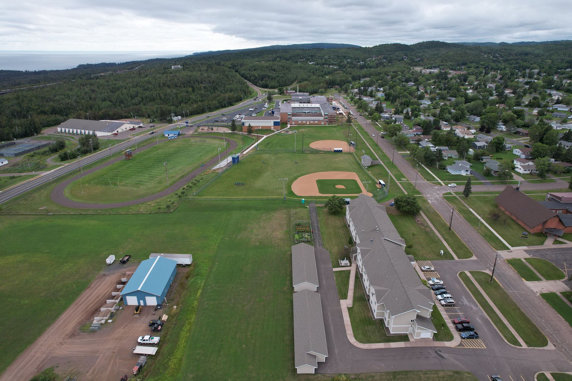 Aerial view of a school complex with sports fields, buildings, and a residential area under a cloudy sky.