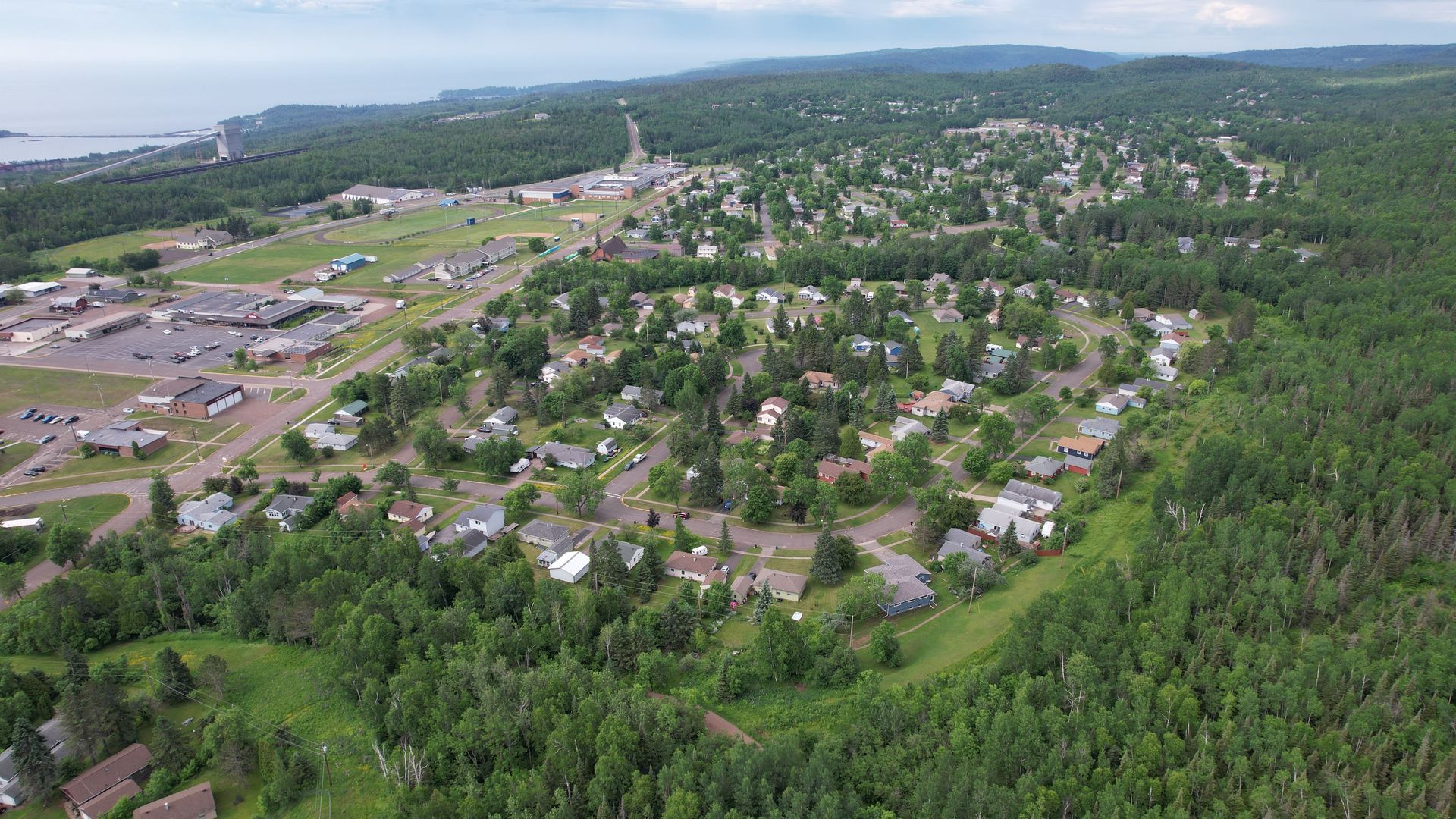 Aerial view of a town near a lake, surrounded by green trees and buildings. Cloudy sky overhead.