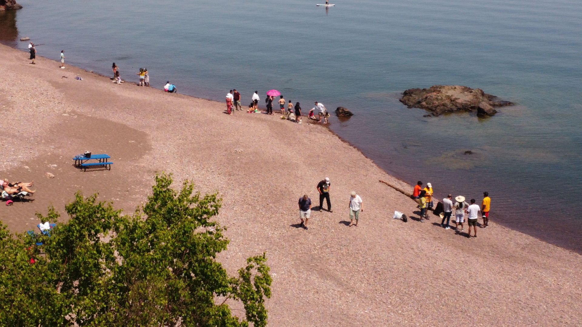 People on a pebbled beach near a lake. Some stand, some bend over. Sunlight, calm water.
