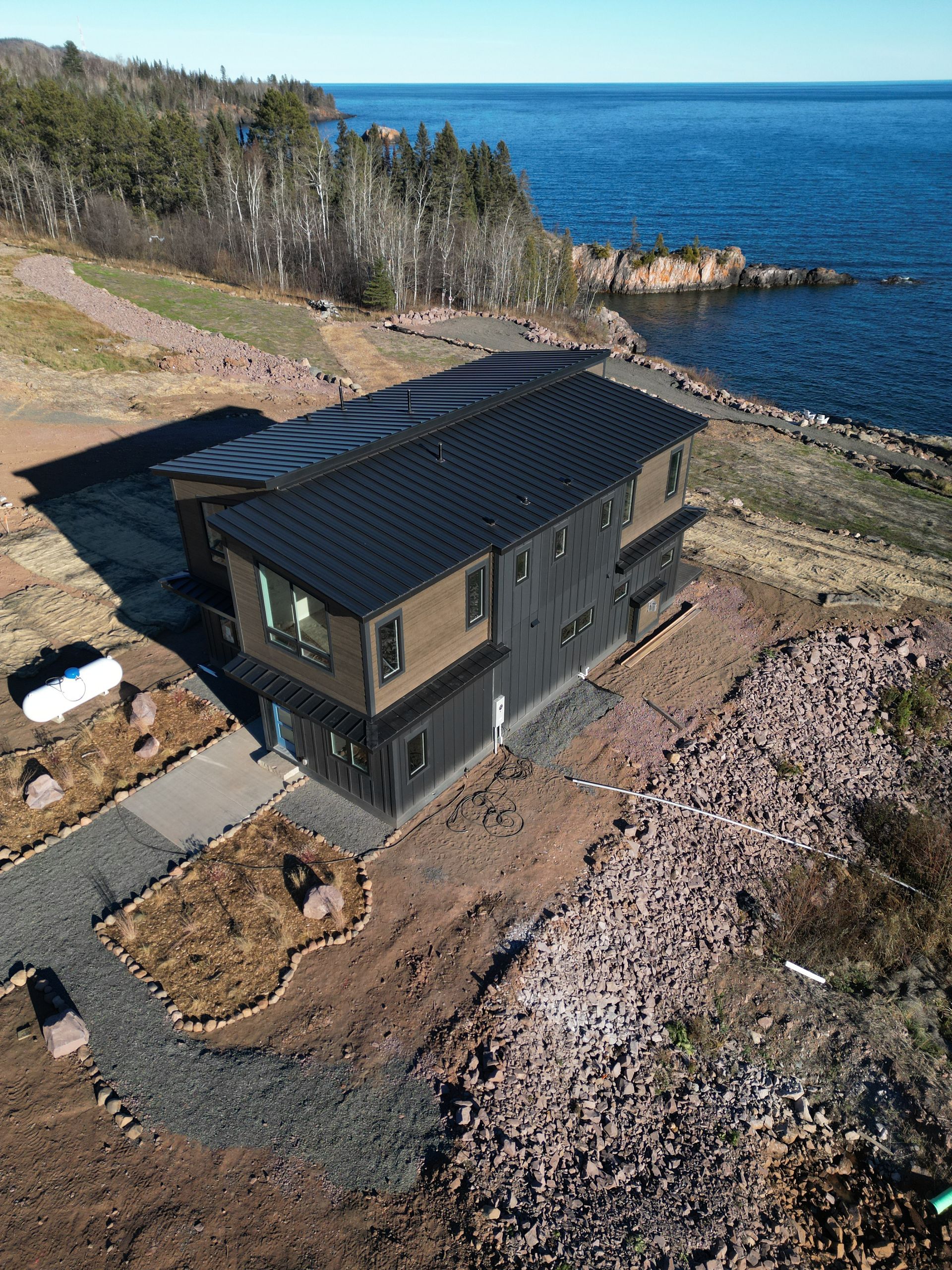 Modern two-story house with dark siding and roof on a rocky coastline, with the blue ocean in the background.