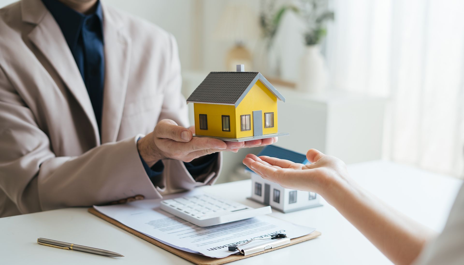 Person handing a yellow toy house to another person, suggesting a real estate transaction.