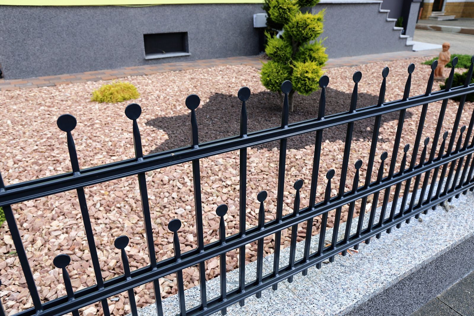Black metal fence with decorative spheres, gravel, and plants in a yard.