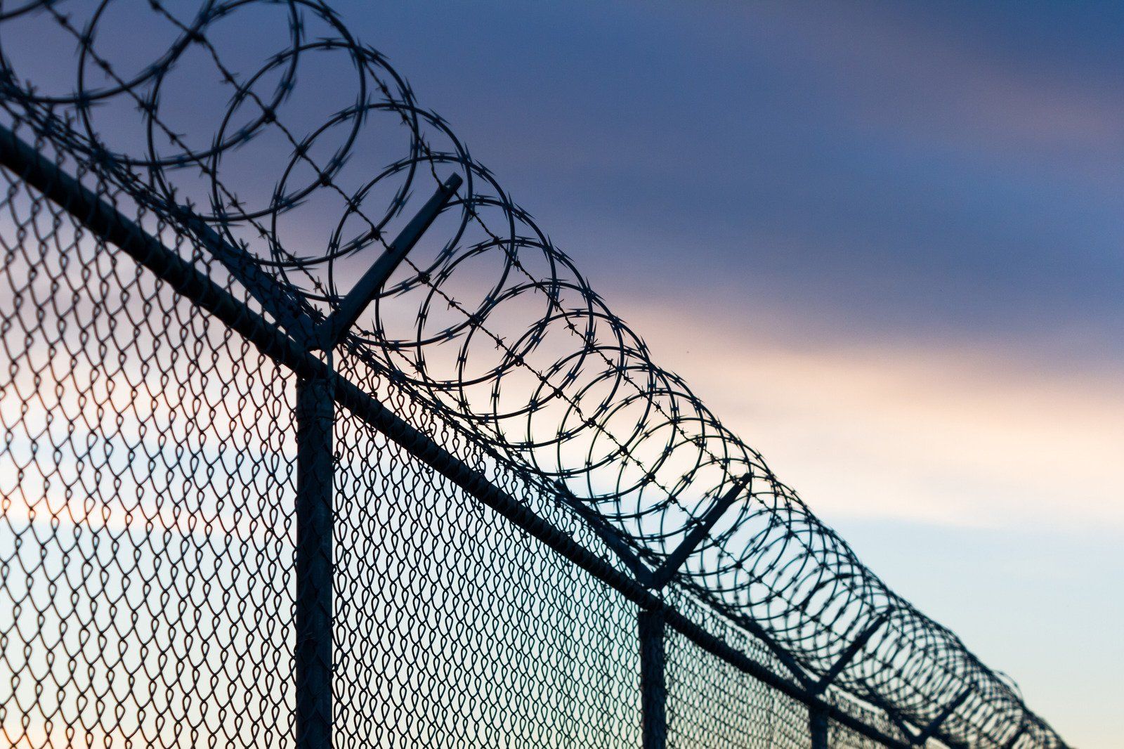 Barbed wire atop chain-link fence against a dusk sky.