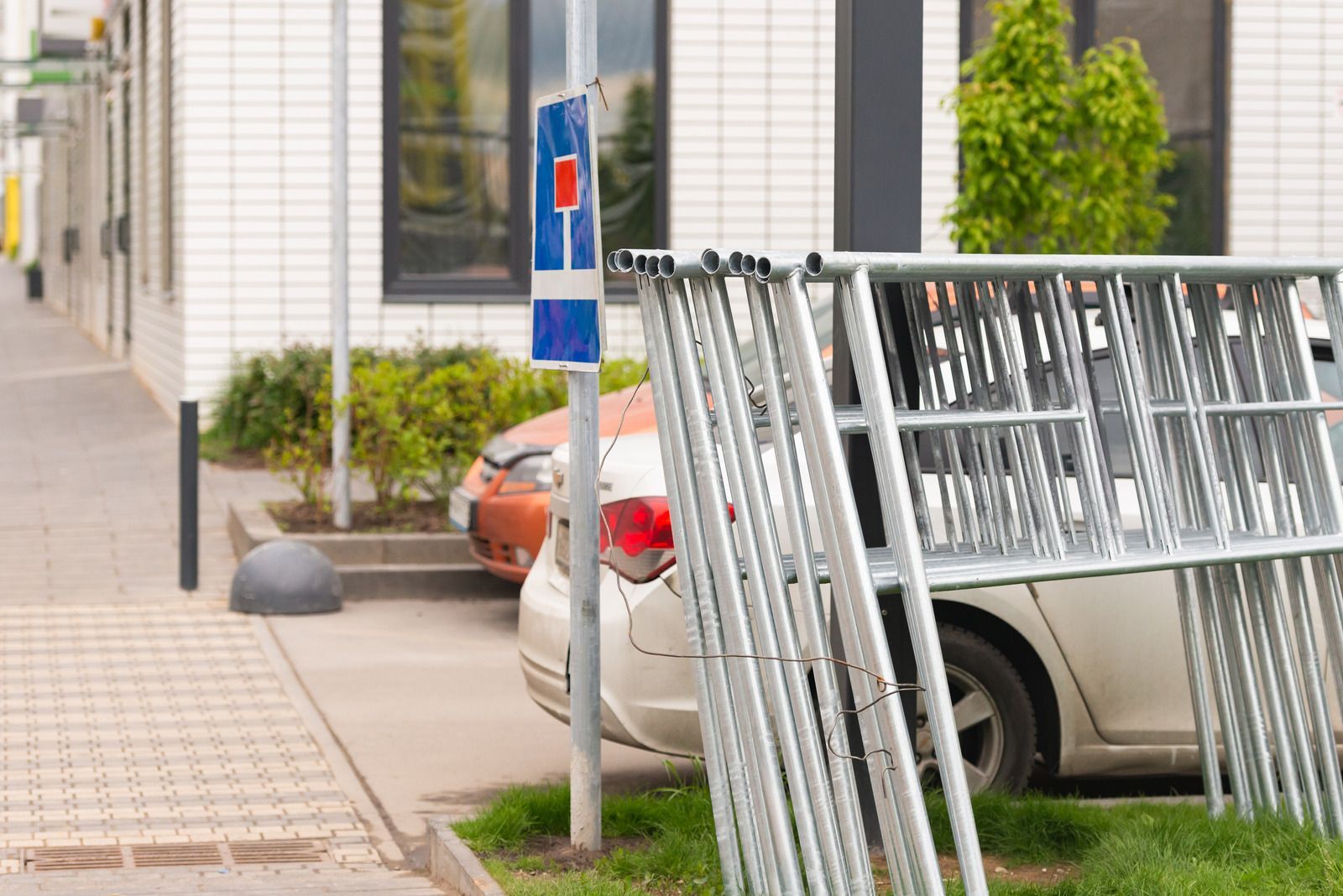 Metal barriers leaning on grass, next to a sign and parked cars near a building.