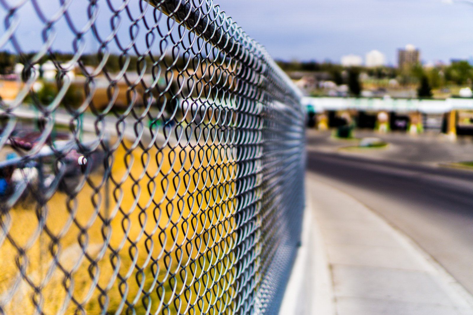 Chain-link fence in focus, with a blurred background of road, buildings, and sky.