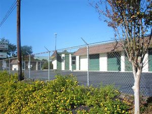 Storage units behind a chain-link fence, with a sign visible on the left. Green doors and a brown roof.