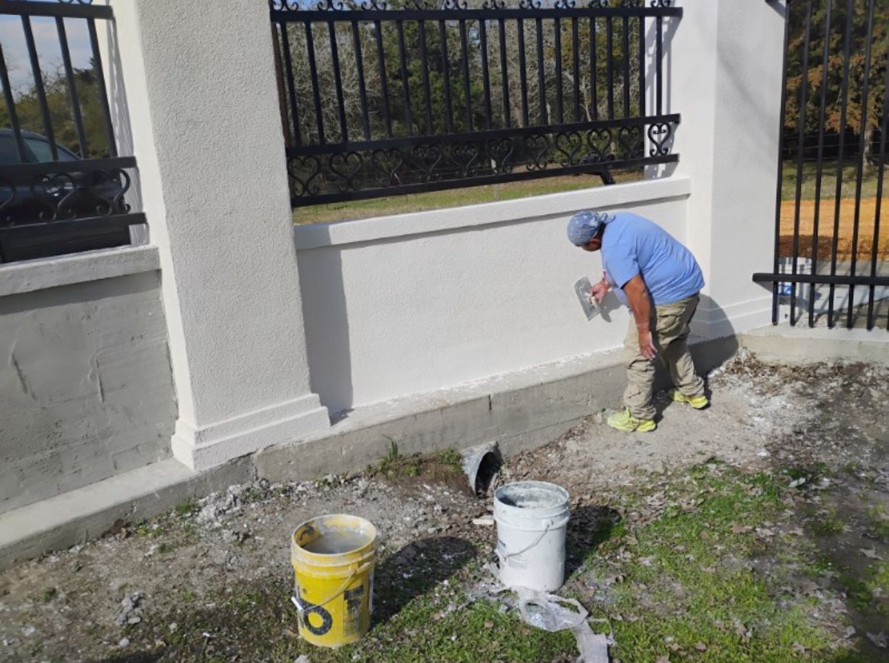 A worker applies stucco to a wall with a trowel outdoors near a black fence and buckets.