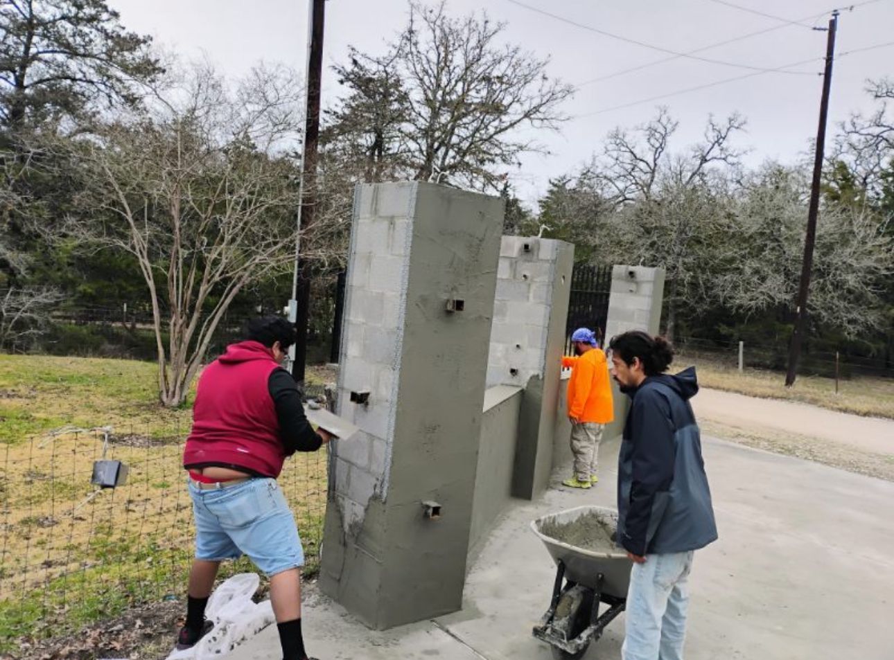 A man is pushing a wheelbarrow full of cement while two other men work on concrete pillars.