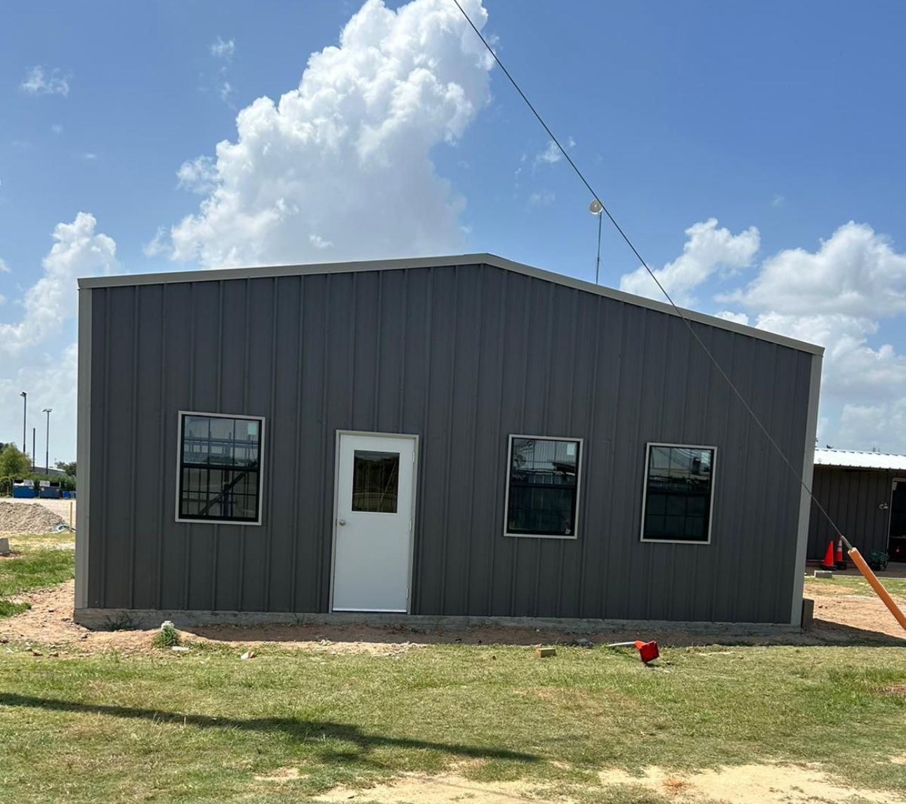 A gray metal building with a white door and windows is sitting in the middle of a grassy field.