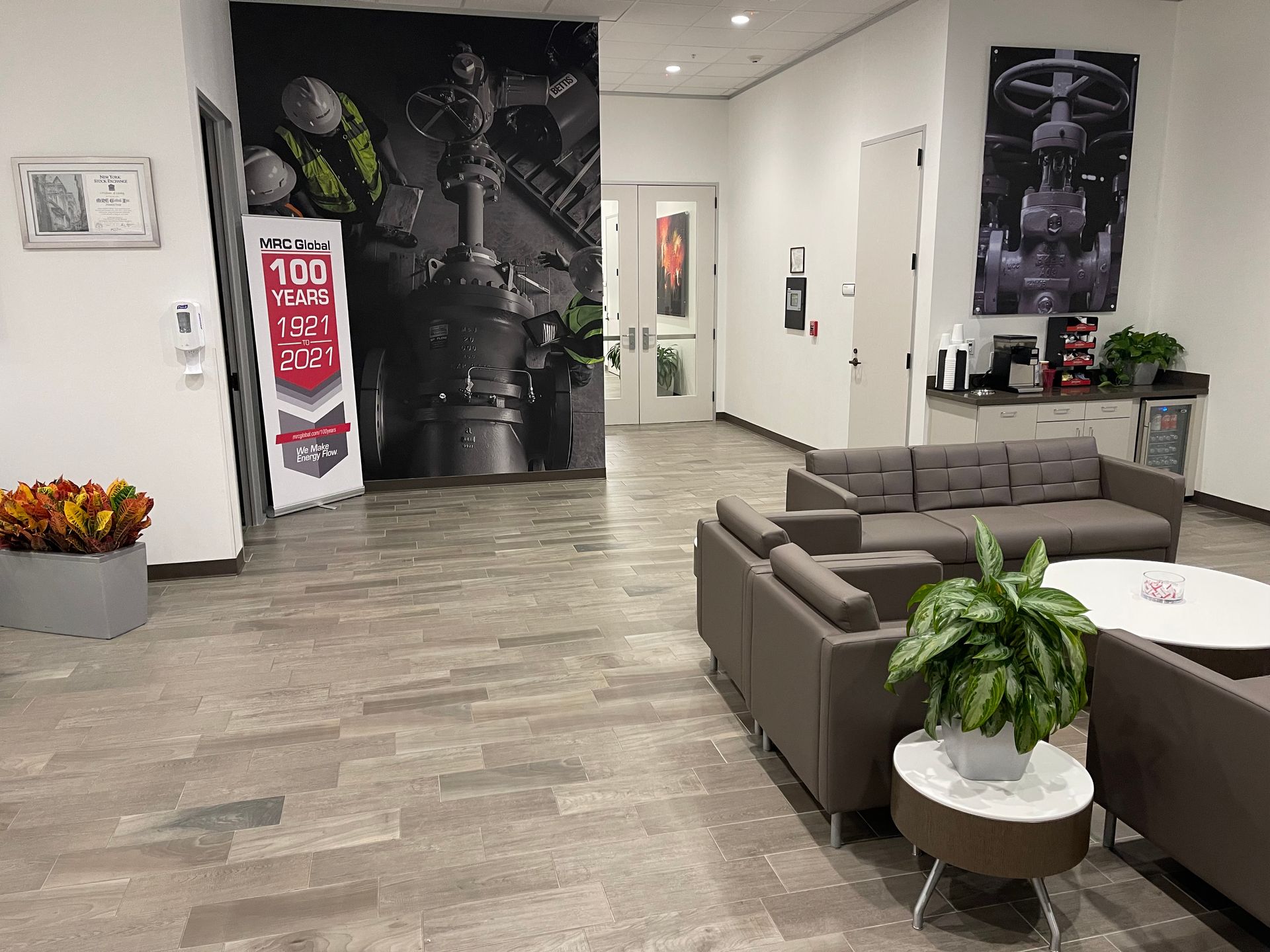 Modern office waiting area with gray couches, potted plants, and a coffee station. A large banner and artwork adorn the walls.