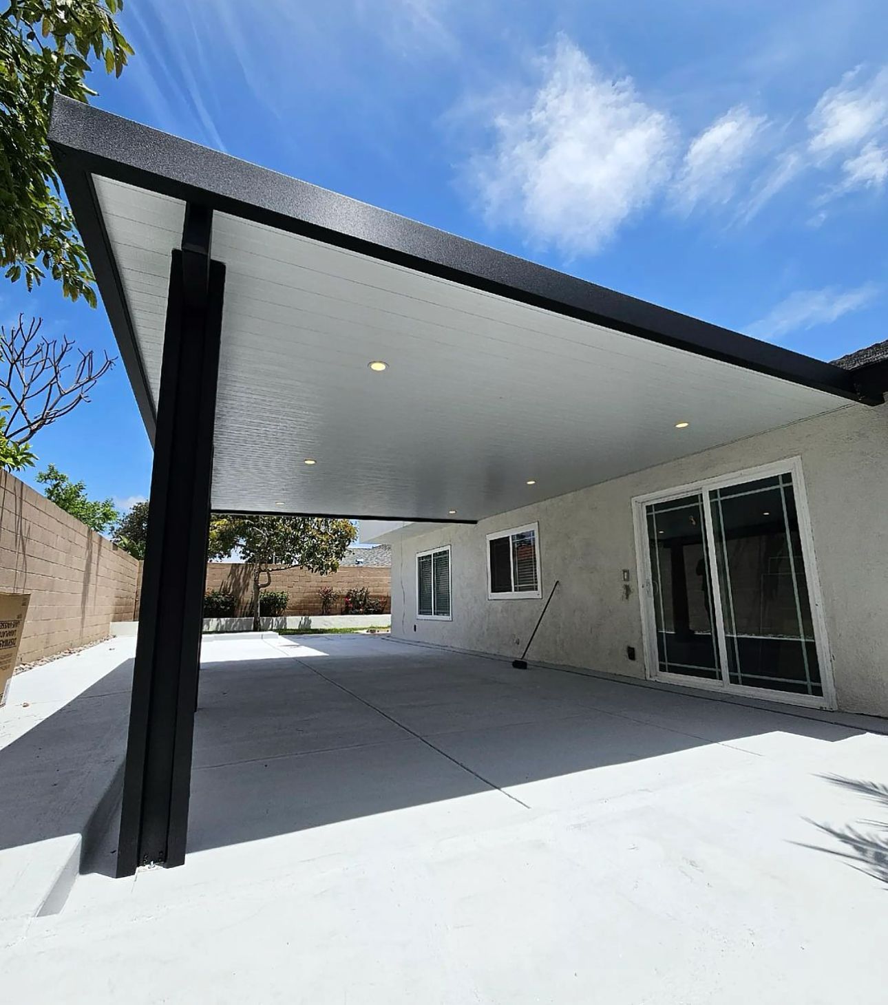 A modern patio cover with black supports, white ceiling, and recessed lights. The patio overlooks a stamped concrete yard with a house and blue sky.