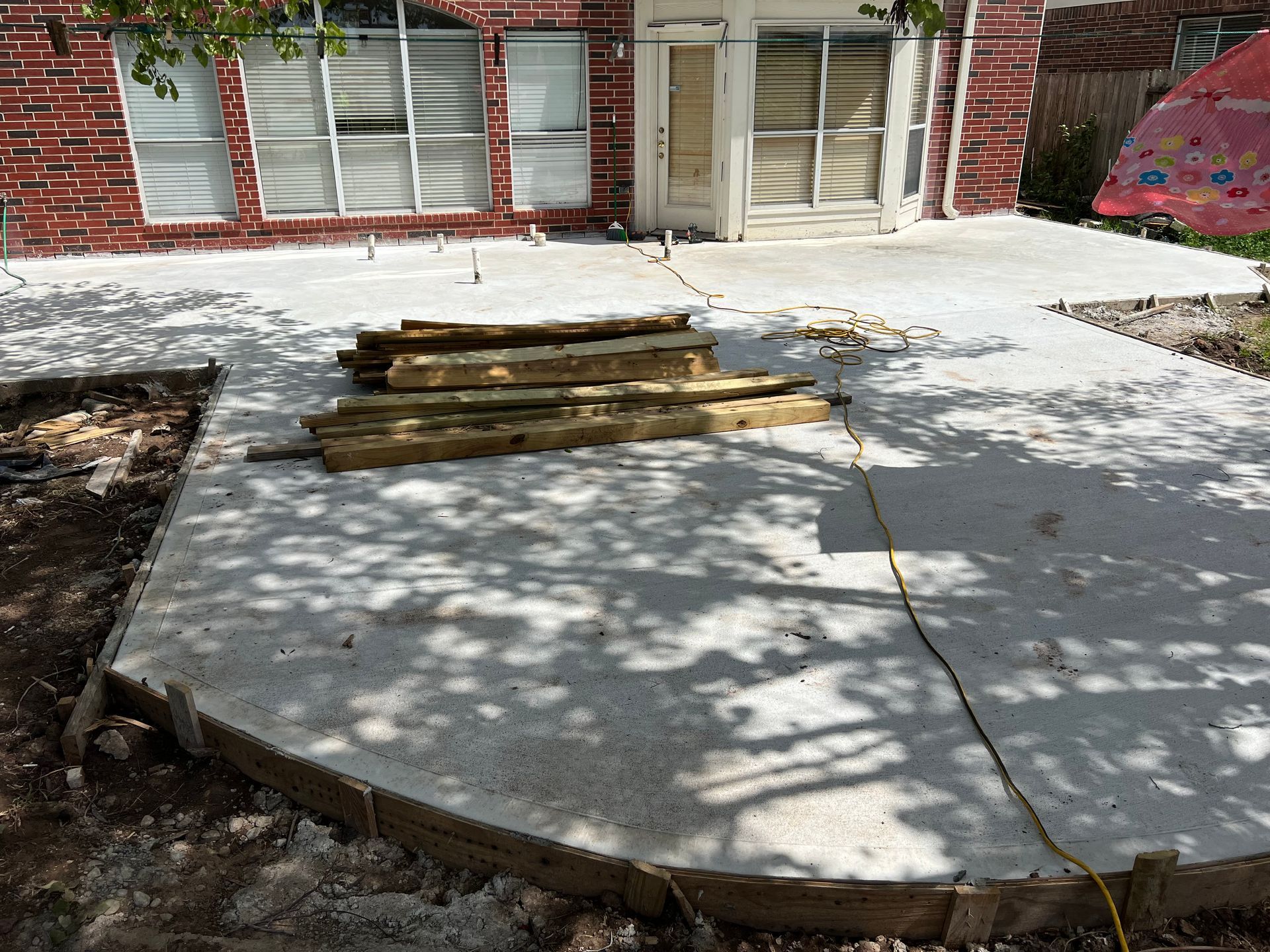 Freshly poured concrete patio with wooden planks on it, set in front of a red brick house.
