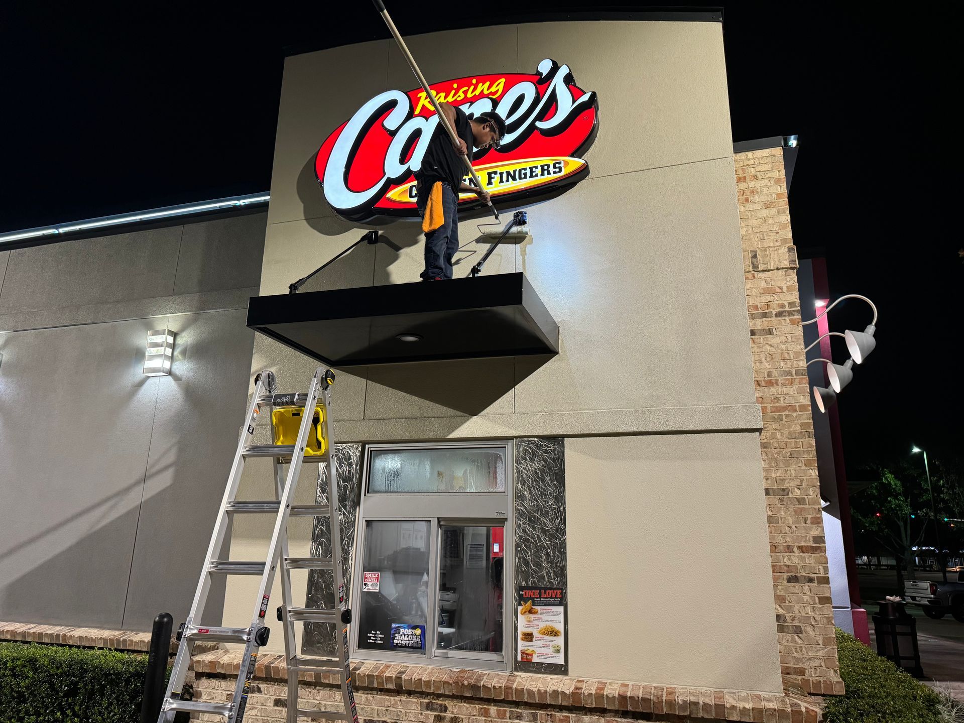 A person cleans the Raising Cane's sign above a drive-thru window at night. A ladder is set up.