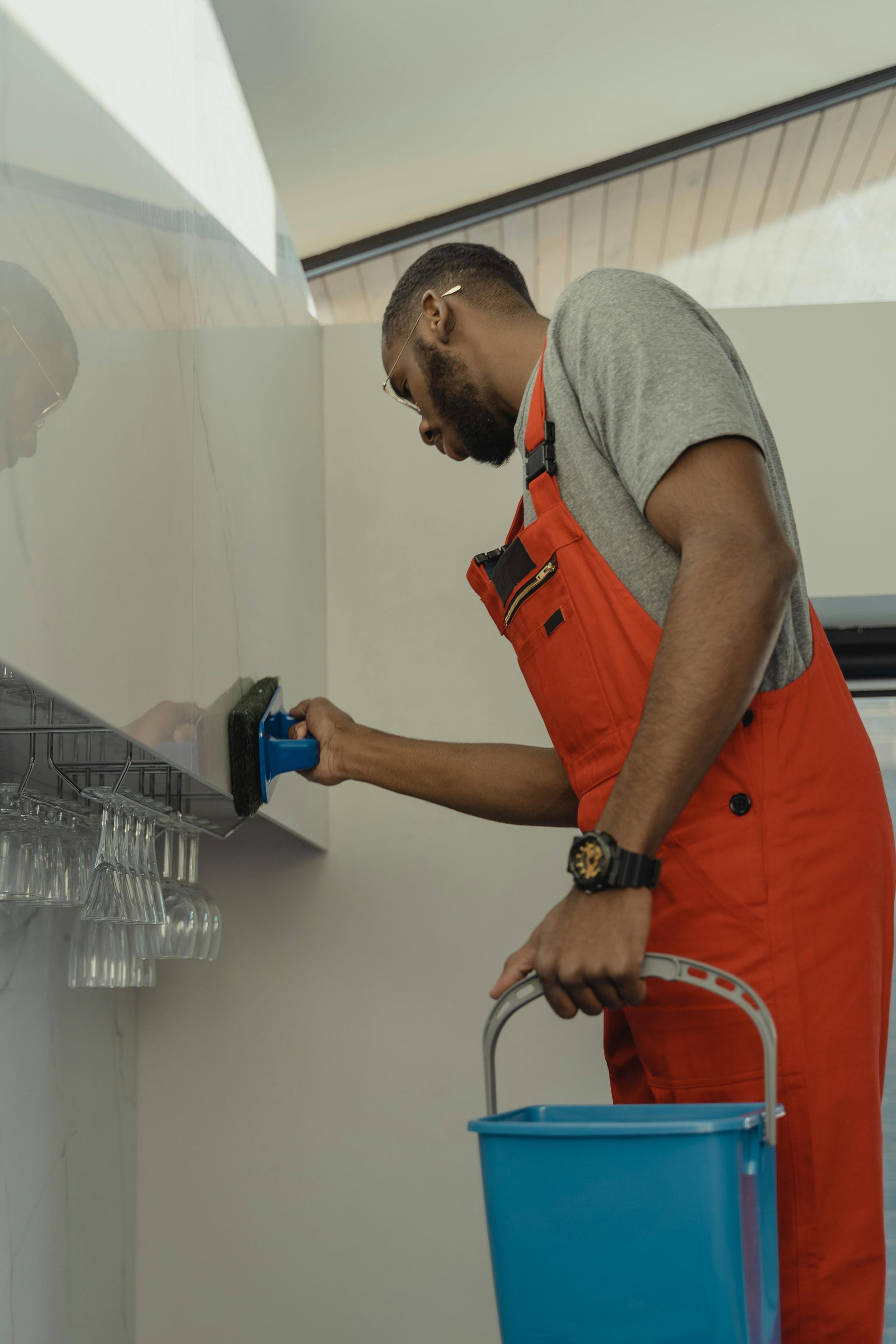 A Black man in orange overalls cleans a wall with a blue bucket at his feet. He appears to be focusing on his task.