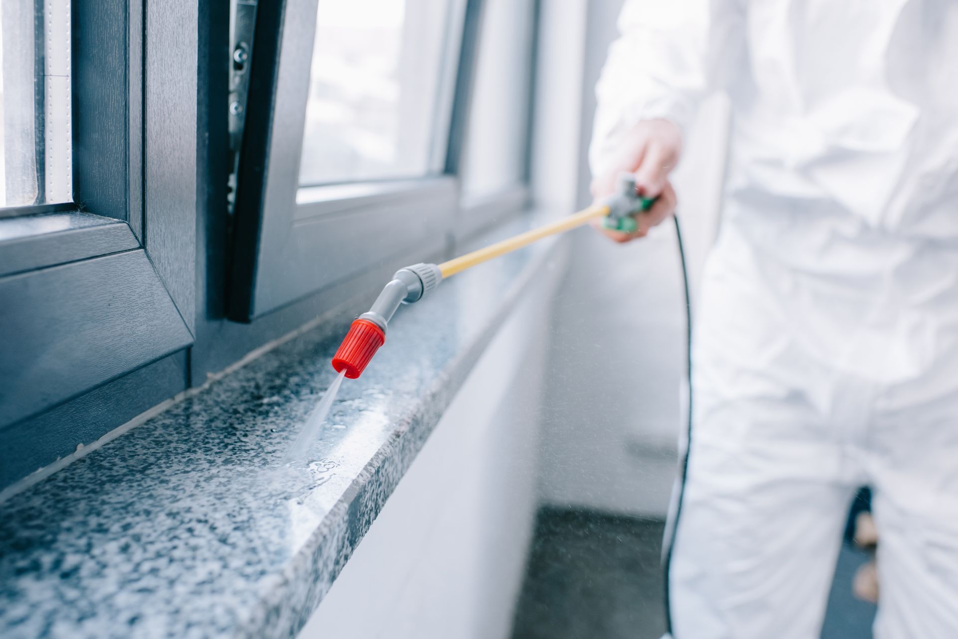 A man in a white suit is spraying a window sill with a sprayer.