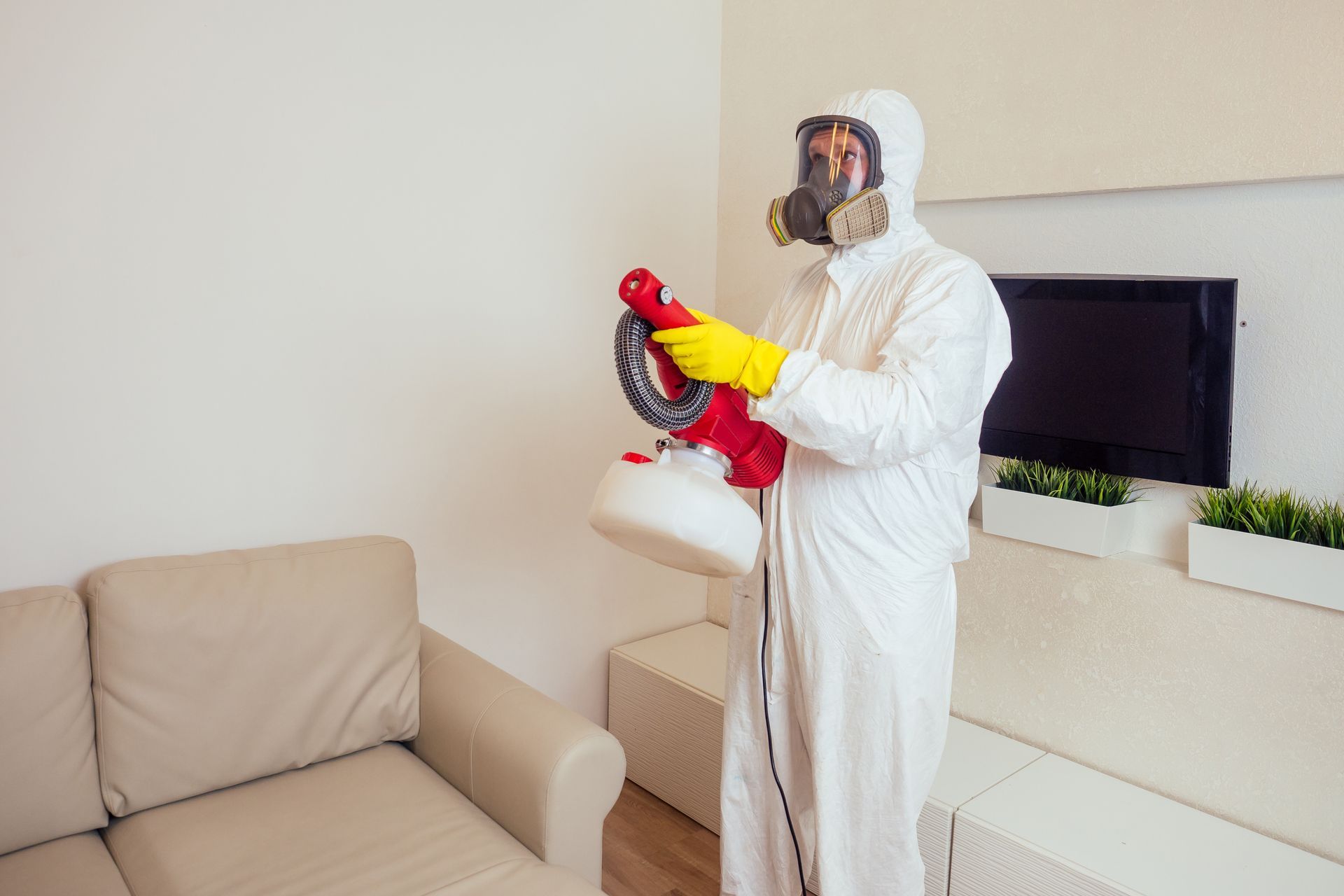 A man in a protective suit and gas mask is disinfecting a living room.