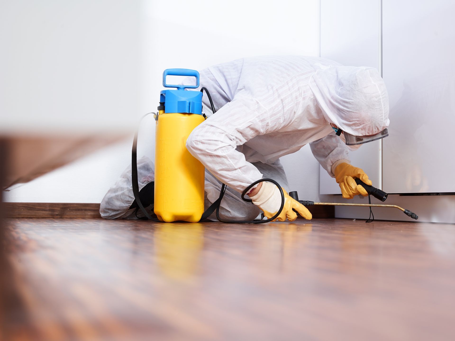 A man in a protective suit is spraying a wooden floor with a sprayer.