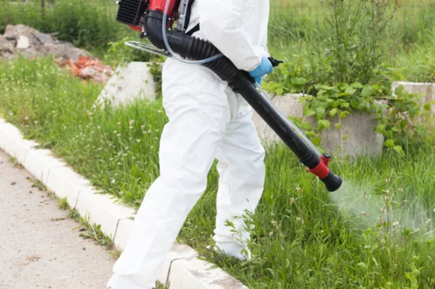 A man in a protective suit is spraying grass with a backpack sprayer.