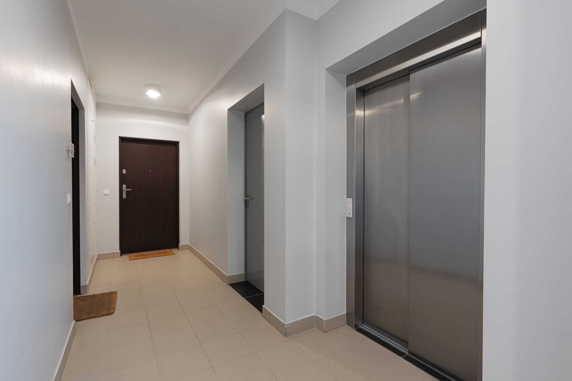 Hallway with a dark door, gray walls, elevator, and a beige tiled floor. Hallway with a dark door, gray walls, elevator, and a beige tiled floor.