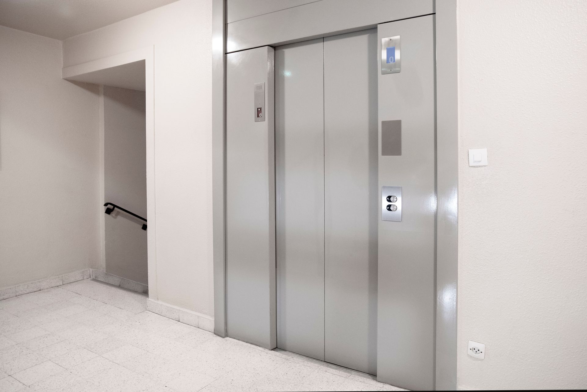 Elevator with closed metal doors in a white-walled hallway. A small button panel is visible. Elevator with closed metal doors in a white-walled hallway. A small button panel is visible.