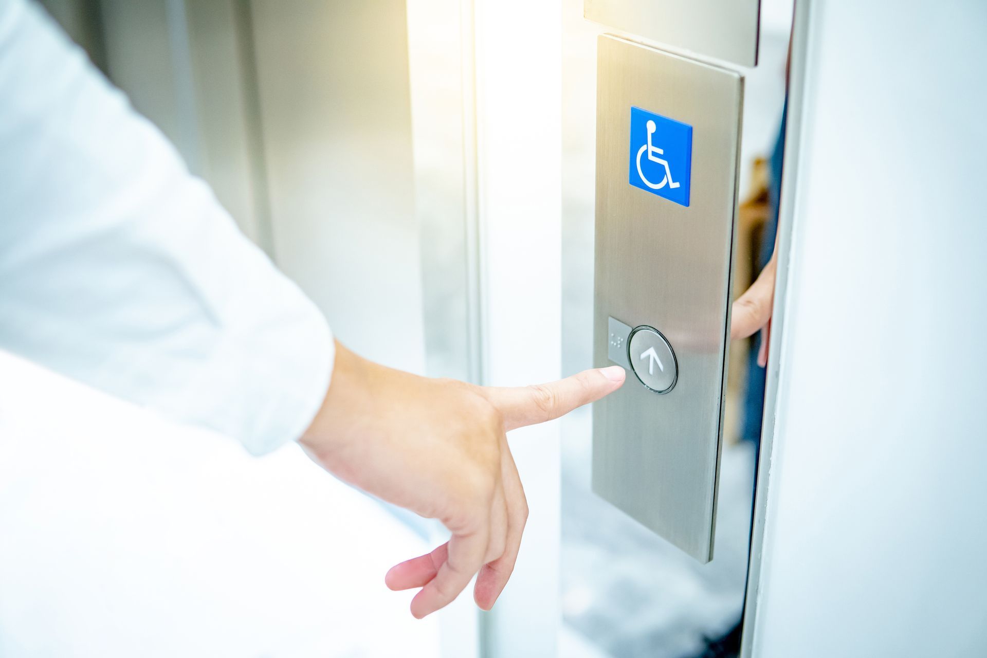 Person's finger pressing an elevator button with accessibility symbol. Silver metal panel. Bright light.
