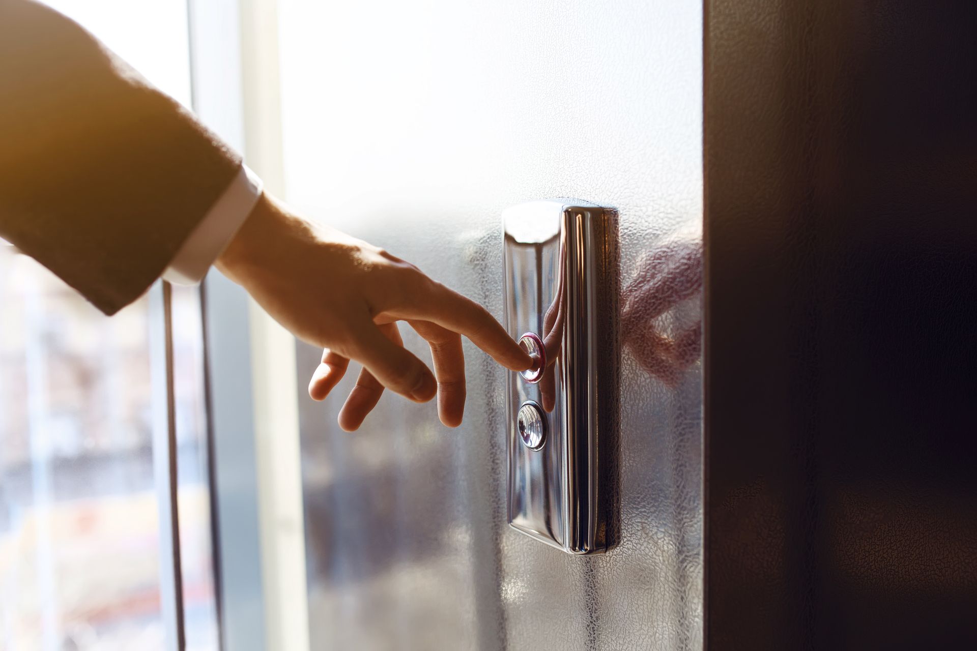 Hand pressing a silver elevator button next to a window. Sunlight illuminates the scene.