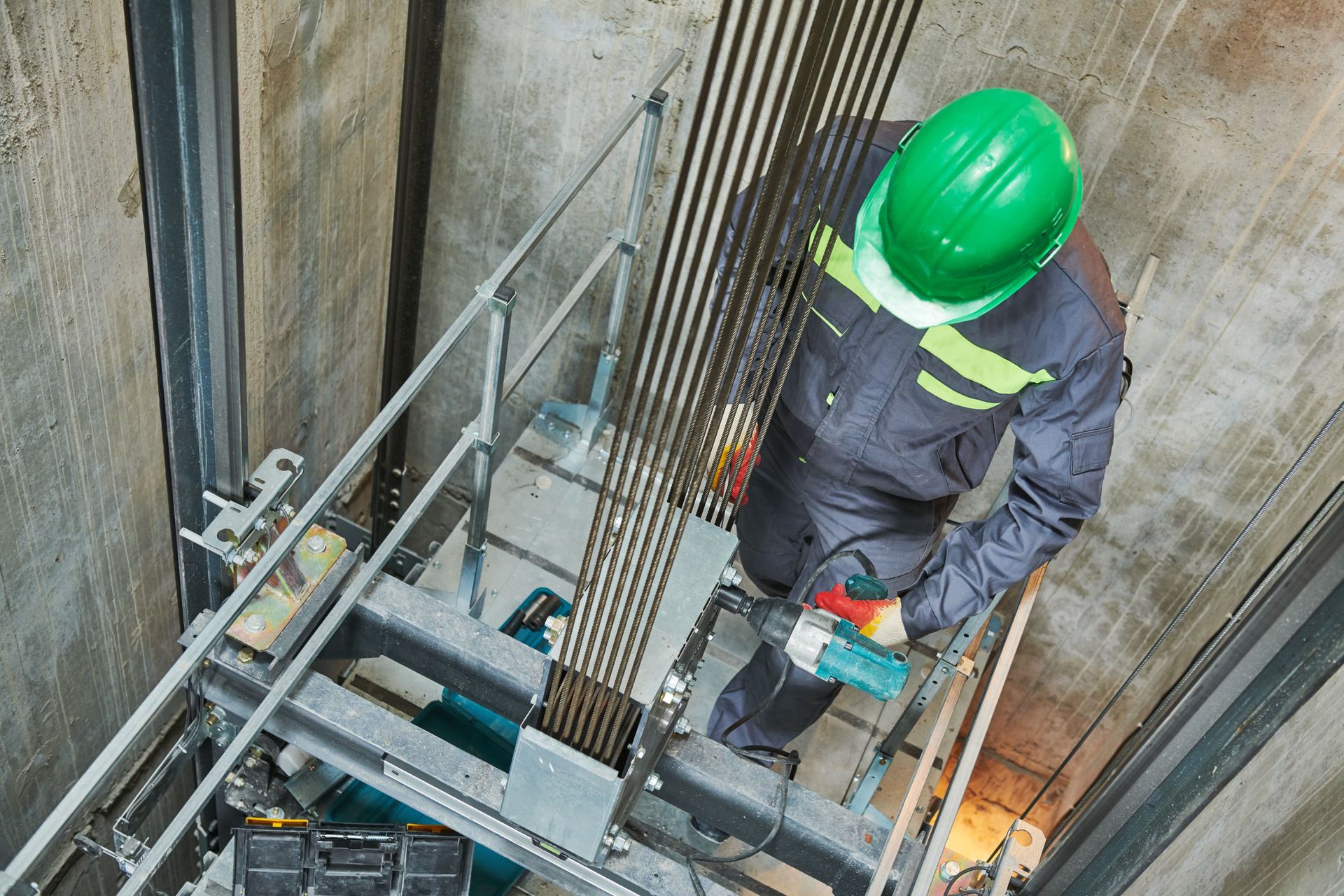 Elevator mechanic in green hard hat, inspecting cables inside an elevator shaft, holding a power tool.