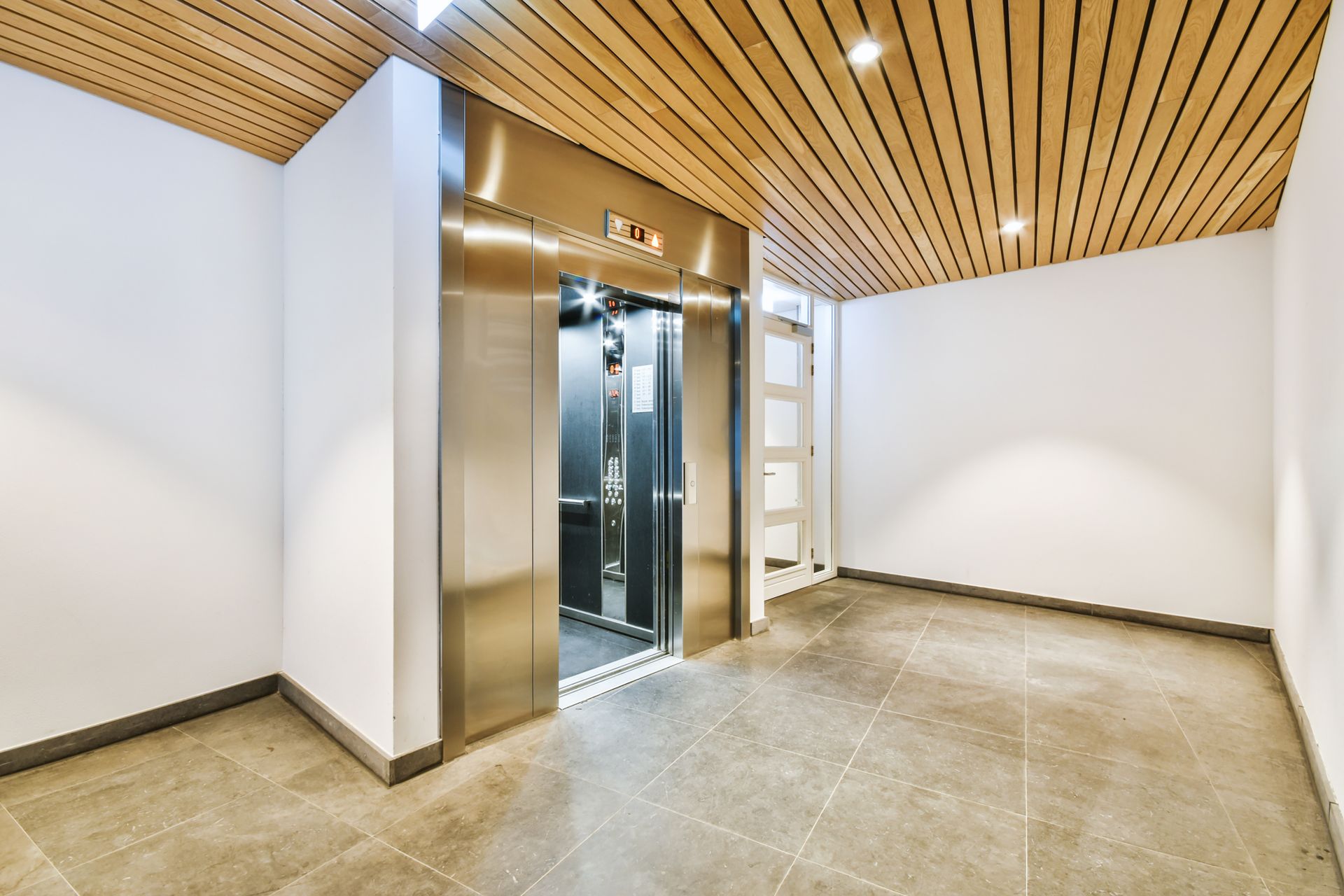 Hallway with elevator. Stainless steel doors, wood-paneled ceiling, gray tile floor, white walls.