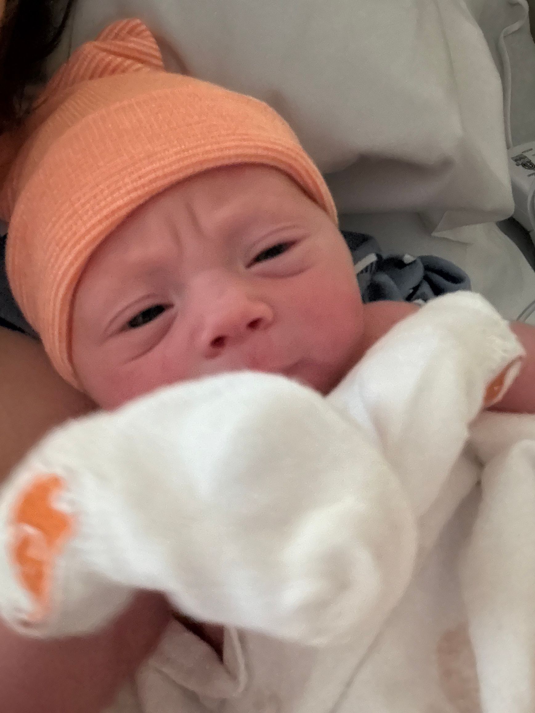 Newborn wearing orange hat, holding white cloth, making a face.