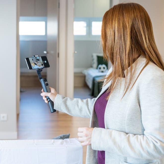 A woman is holding a phone on a tripod in a room recording a selfie video.
