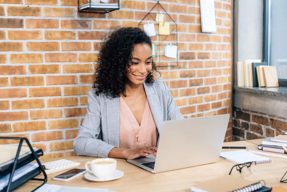 Woman typing on laptop at wooden desk near brick wall. Cup of coffee and papers on desk.