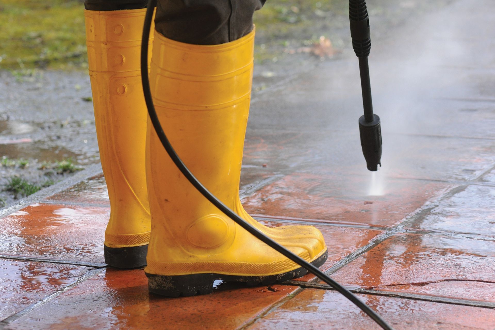 A person wearing yellow boots is using a high pressure washer to clean a sidewalk.