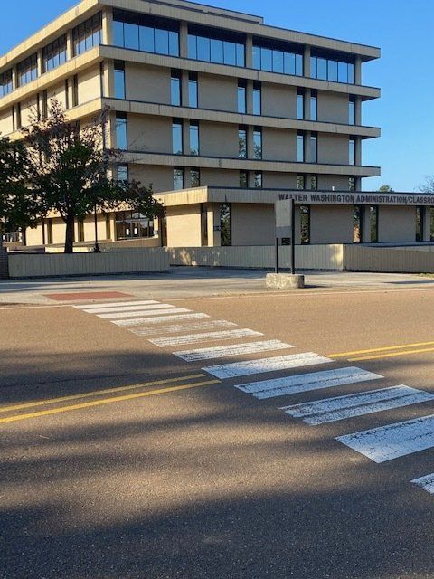 Crosswalk in front of a beige, multi-story building with a sign; sunny day.